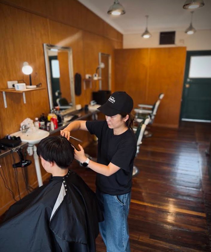 A Tokoya barber in a black cap cutting a customer's hair with scissors.