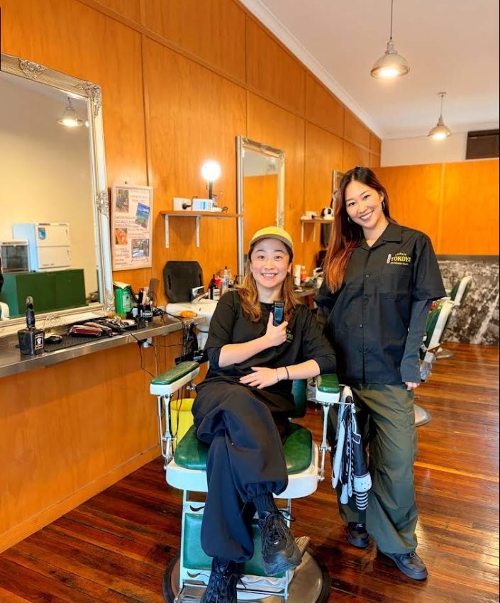 Two smiling Tokoya barbers beside a green leather barber chair.